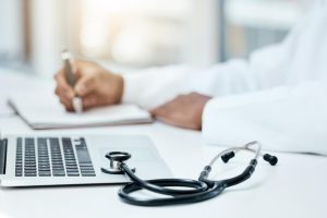 Photograph of a doctor sitting at a desk, writing; only his arms are visible. In front of him is a laptop, and next to the laptop is a stethoscope.