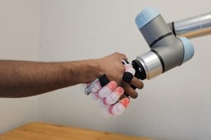 A robotic arm and a human hand engaged in a handshake, with the robotic system displaying soft and hard materials.